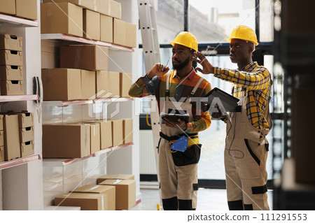 Warehouse manager training assistant doing inventory with barcode scanner and tablet app. Young african american storehouse employee taking notes while supervisor explaining stock management Warehouse manager training assistant doing inventory with barcode scanner and tablet app. Young african american storehouse employee taking notes while supervisor explaining stock management 111291553