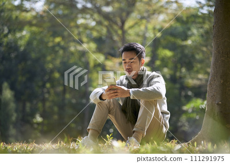 young asian adult man sitting on grass in park looking at cellphone young asian adult man sitting on grass in park looking at cellphone 111291975