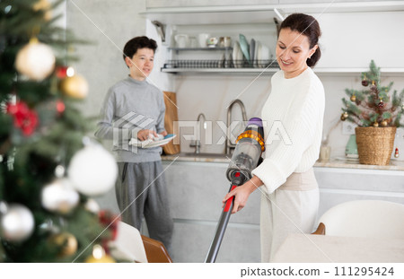 Preparing for Christmas - mother vacuuming floor while a teenage son washes dishes in kitchen 111295424