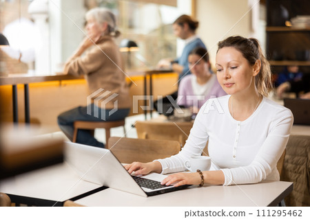 Smiling woman using laptop and drinking coffee in modern cafe 111295462