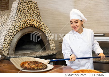 Portrait of young woman chef working in restaurant kitchen, getting ready pizza out of oven Portrait of young woman chef working in restaurant kitchen, getting ready pizza out of oven 111296079