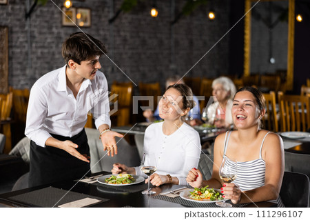 Two young women enjoying lunch in cozy restaurant, where waiter attentively serves them. Two young women enjoying lunch in cozy restaurant, where waiter attentively serves them. 111296107
