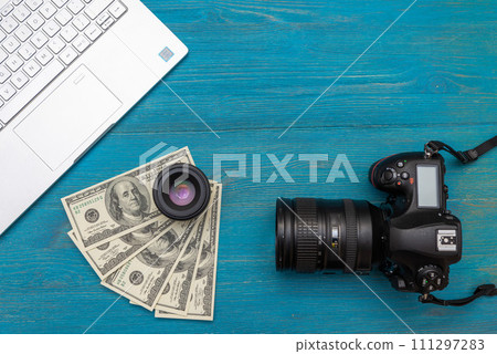 Earn money in internet DSLR camera, dollars, laptop, on blue wooden background, top view 111297283