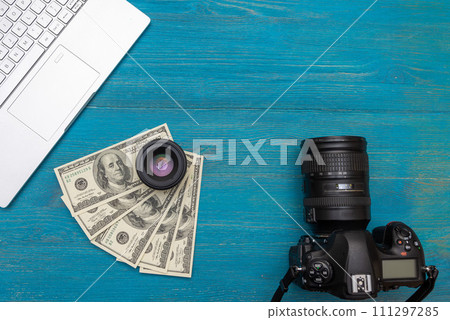 Earn money in internet DSLR camera, dollars, laptop, on blue wooden background, top view Earn money in internet DSLR camera, dollars, laptop, on blue wooden background, top view 111297285