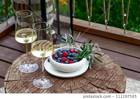 Two glasses of white wine and a plate of wild blueberries and raspberries on a wooden table in a cafe. High angle view. Two glasses of white wine and a plate of wild blueberries and raspberries on a wooden table in a cafe. High angle view. 111297585