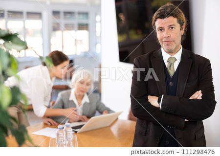 positive male colleague standing in meeting room and employees are talking behind him at table 111298476