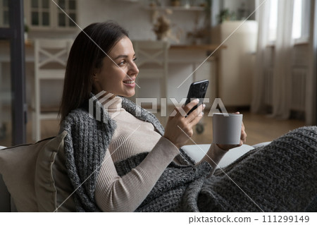 Woman covered with plaid hold teacup, smartphone relaxing at home Woman covered with plaid hold teacup, smartphone relaxing at home 111299149