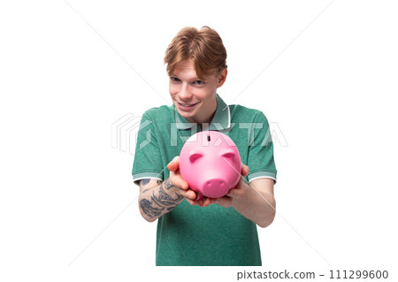 a young European man with red hair holds out a pink piggy bank with money and coins a young European man with red hair holds out a pink piggy bank with money and coins 111299600