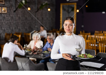 Young female waitress stands with order tray in spacious guest room in restaurant. Young female waitress stands with order tray in spacious guest room in restaurant. 111299830