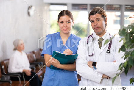 Young woman and adult man doctors posing in clinic reception Young woman and adult man doctors posing in clinic reception 111299863