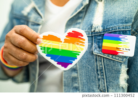 Asian lady wearing rainbow flag wristbands and hold red heart, symbol of LGBT pride month celebrate annual in June social of gay, lesbian, bisexual, transgender, human rights. Asian lady wearing rainbow flag wristbands and hold red heart, symbol of LGBT pride month celebrate annual in June social of gay, lesbian, bisexual, transgender, human rights. 111300452