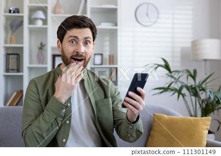 Stunned caucasian guy holding smartphone in hand and cover mouth with hand while looking at camera with amazement. Impressed man demonstrating reaction for unexpected text message on mobile device. Stunned caucasian guy holding smartphone in hand and cover mouth with hand while looking at camera with amazement. Impressed man demonstrating reaction for unexpected text message on mobile device. 111301149