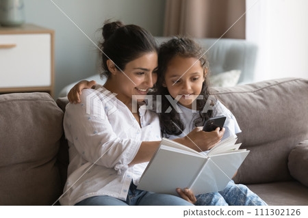 Mother and daughter sit on sofa holding book using smartphone Mother and daughter sit on sofa holding book using smartphone 111302126
