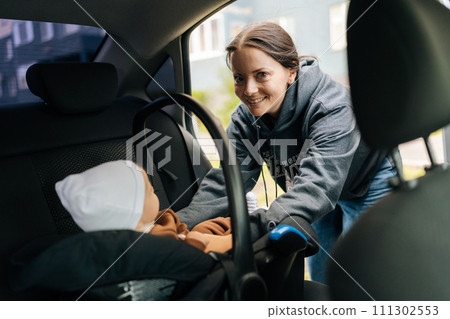 Portrait of positive pretty young mom placing child safety seat with infant baby boy on back passenger seat, smiling looking at camera, view from front passenger seat. Concept of safety lifestyle. 111302553