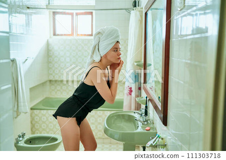 Woman in bathroom, towel on head, treats facial acne, removes pimples. Mirror reflection shows skincare routine, imperfection care, personal hygiene, beauty regimen, and dermatology at home. Woman in bathroom, towel on head, treats facial acne, removes pimples. Mirror reflection shows skincare routine, imperfection care, personal hygiene, beauty regimen, and dermatology at home. 111303718