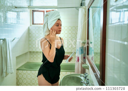 Woman in retro bathroom applies moisturizer to face. Skincare routine with cream for beauty, health. Female in towel maintains complexion, uses cosmetic product. Vintage interior echoes self-care. Woman in retro bathroom applies moisturizer to face. Skincare routine with cream for beauty, health. Female in towel maintains complexion, uses cosmetic product. Vintage interior echoes self-care. 111303728