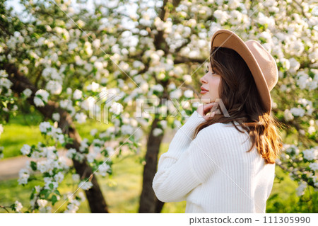 Portrait of Beautiful Woman In hat posing near flowering. Smiling woman enjoying smell of flowers. Portrait of Beautiful Woman In hat posing near flowering. Smiling woman enjoying smell of flowers. 111305990