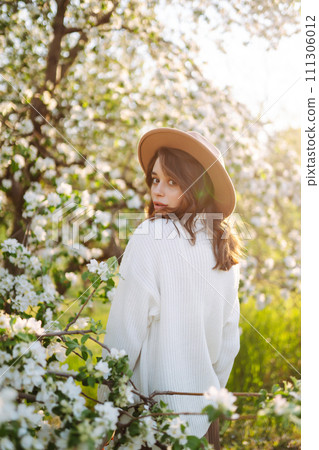 Portrait of Beautiful Woman In hat posing near flowering. Smiling woman enjoying smell of flowers. Portrait of Beautiful Woman In hat posing near flowering. Smiling woman enjoying smell of flowers. 111306012