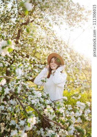 Portrait of Beautiful Woman In hat posing near flowering. Smiling woman enjoying smell of flowers. 111306015