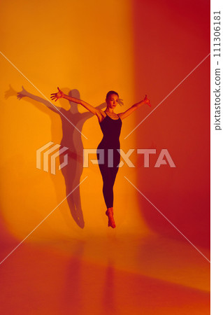 Athletic Grace. Body-Size Portrait of beautiful woman, ballerina dancer stretches in black sports overalls, barefoot in neon-lit studio. Athletic Grace. Body-Size Portrait of beautiful woman, ballerina dancer stretches in black sports overalls, barefoot in neon-lit studio. 111306181