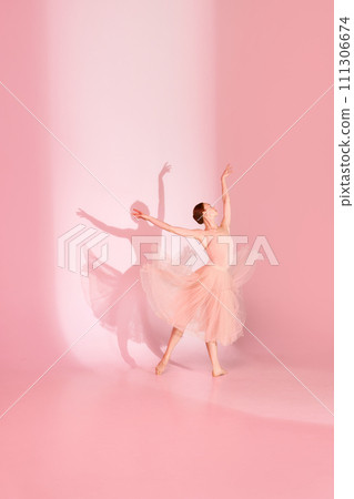 Graceful Ballet. Young woman, classic ballerina dancing barefoot in pink dress against pastel studio background with her shadow. Graceful Ballet. Young woman, classic ballerina dancing barefoot in pink dress against pastel studio background with her shadow. 111306674