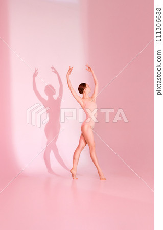 Pastel Ballet. Woman gracefully poses in pink swimsuit against soft pink backdrop. Her barefoot silhouette casts shadow, echoing her elegant movements. 111306688
