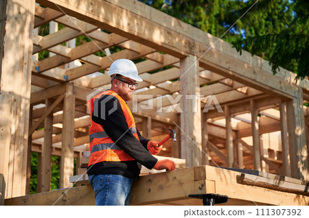 Carpenter constructing wooden frame house near forest. Bearded man with glasses hammering nails into structure, wearing protective helmet, construction vest. Concept of modern ecological construction. 111307392