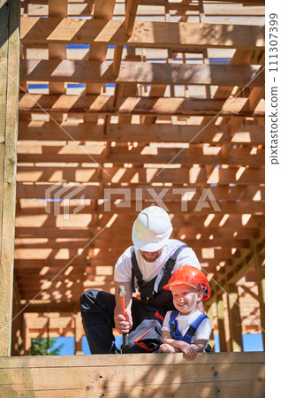 Father with toddler son building wooden frame house. Male builder teaching his happy son the technique of pounding nails with hammer on construction site, wearing helmets and overalls on sunny day. 111307399