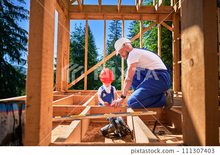 Father with toddler son building wooden frame house. Male builder playing with kid on construction site, wearing helmets and blue overalls on sunny day. Carpentry and family concept. 111307403