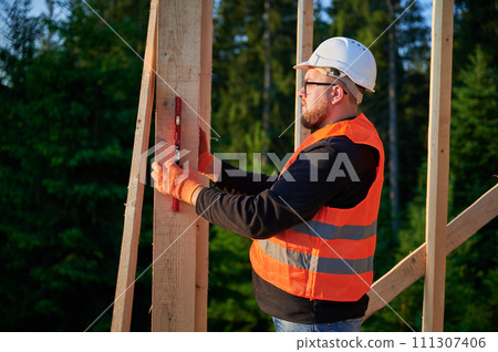 Carpenter constructing wooden frame house near the forest. Bearded man, wearing spectacles, inspects the walls for levelness using spirit level. He is attired in protective helmet, and orange vest. Carpenter constructing wooden frame house near the forest. Bearded man, wearing spectacles, inspects the walls for levelness using spirit level. He is attired in protective helmet, and orange vest. 111307406