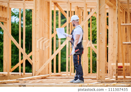 Construction worker building two-story wooden frame house. Man with beard, wearing jumpsuit and hard hat, reviewing the building plan. Intention to implement modern ecological construction practices. Construction worker building two-story wooden frame house. Man with beard, wearing jumpsuit and hard hat, reviewing the building plan. Intention to implement modern ecological construction practices. 111307435