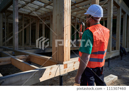 Carpenter constructing two-story wooden frame house near the forest. Back view of man hammering nails into structure while wearing protective helmet and construction vest. 111307460