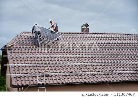 Men technicians carrying photovoltaic solar modul on roof of house. Builders in helmets building solar panel system outdoors. Concept of alternative and renewable energy. 111307467