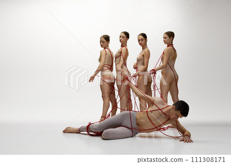Ballet art. Making choice. Male dancer lies on floor while four female dancers stand near, all connected by red strings against white studio background Ballet art. Making choice. Male dancer lies on floor while four female dancers stand near, all connected by red strings against white studio background 111308171
