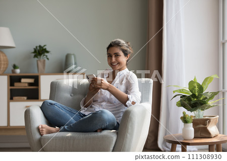 Attractive young Indian woman relaxing on cosy armchair with smartphone Attractive young Indian woman relaxing on cosy armchair with smartphone 111308930