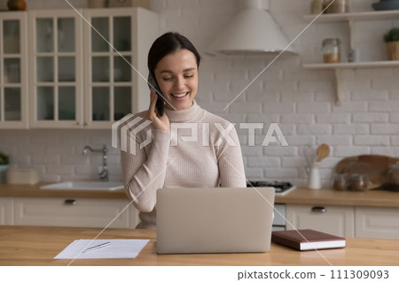 Attractive woman sit in kitchen with laptop talking on cellphone 111309093