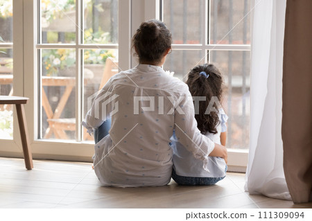 Rear view mother hugs little daughter looking out window together 111309094