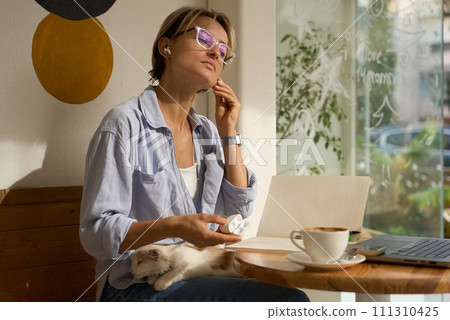 Young smiling woman in cafe with cute small cat sitting at table with laptop, female freelancer Young smiling woman in cafe with cute small cat sitting at table with laptop, female freelancer 111310425
