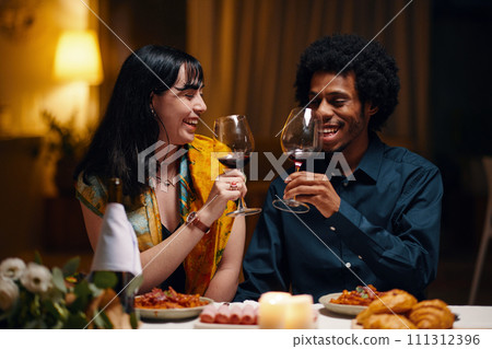 Happy young man and woman with glasses of red wine toasting for love and Valentine Day while sitting by served table at dinner Happy young man and woman with glasses of red wine toasting for love and Valentine Day while sitting by served table at dinner 111312396