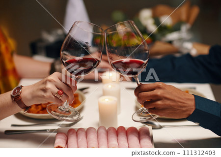 Hands of young unrecognizable sweethearts clinking with glasses of wine over table served with homemade food and fresh platter Hands of young unrecognizable sweethearts clinking with glasses of wine over table served with homemade food and fresh platter 111312403