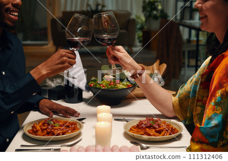Cropped shot of happy young intercultural valentines clinking with glasses of red wine while sitting by served table with homemade food 111312406