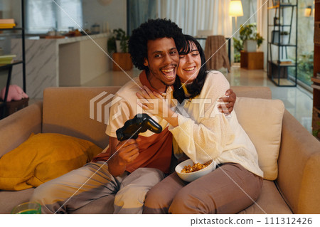 Young intercultural man and woman sitting in embrace on couch in front of TV set during video game while guy looking at screen Young intercultural man and woman sitting in embrace on couch in front of TV set during video game while guy looking at screen 111312426