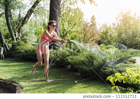 Young adult sexy slim sporty caucasian brunette woman enjoy having fun watering garden at home yard with hose pipe shower on hot summer day. Happy female person relax at backyard park outdoors 111312711