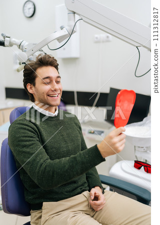 Vertical portrait of handsome cheerful young male patient smiling into mirror looking at healthy white teeth enjoying dental healthcare facilities in modern dentist office. Concept of dentistry. Vertical portrait of handsome cheerful young male patient smiling into mirror looking at healthy white teeth enjoying dental healthcare facilities in modern dentist office. Concept of dentistry. 111312817