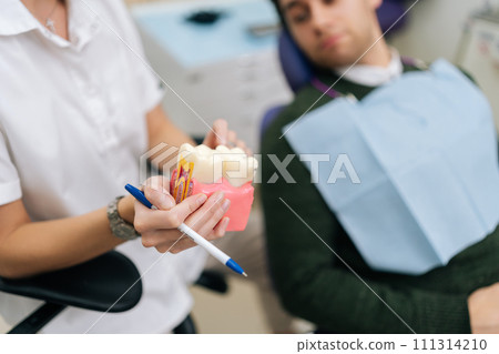 High-angle closeup view of unrecognizable female dentist explaining how to take care oral cavity model and teeth include dental disease to male patient, sitting on dental chair in dentistry clinic. High-angle closeup view of unrecognizable female dentist explaining how to take care oral cavity model and teeth include dental disease to male patient, sitting on dental chair in dentistry clinic. 111314210