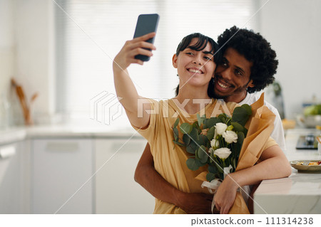 Young cheerful brunette woman holding smartphone while taking selfie by kitchen table with affectionate husband embracing her Young cheerful brunette woman holding smartphone while taking selfie by kitchen table with affectionate husband embracing her 111314238
