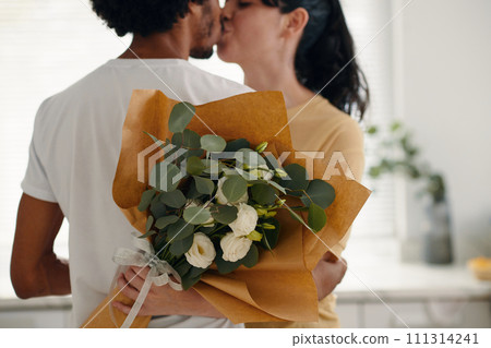 Bunch of beautiful white flowers wrapped in paper held by young amorous woman embracing her husband during passionate kiss 111314241
