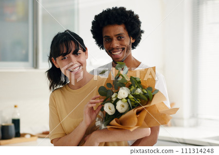 Young cheerful intercultural husband and wife with bunch of white flowers wrapped in paper looking at camera while standing in the kitchen Young cheerful intercultural husband and wife with bunch of white flowers wrapped in paper looking at camera while standing in the kitchen 111314248