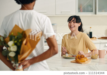 Young cheerful woman in beige t-shirt having breakfast and looking at her husband hiding bunch of white flowers behind back 111314344