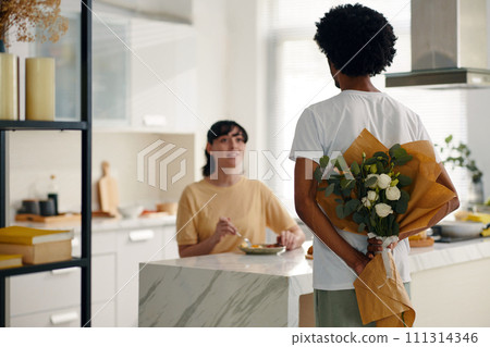 Back of young African American man holding bouquet of white roses for his wife or girlfriend sitting by table in front of him 111314346